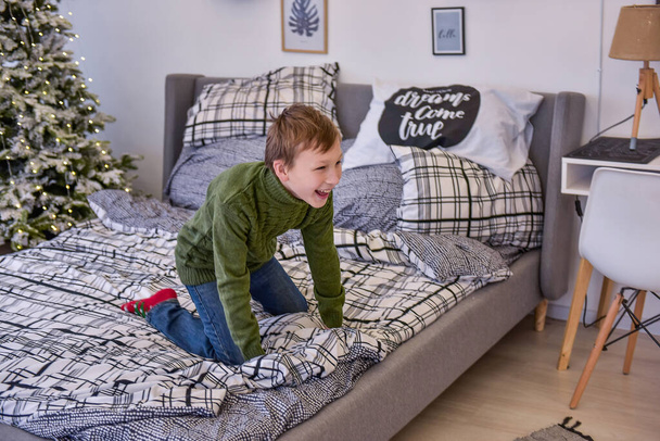 Little boy in green knitted sweater is having fun on stylish bed by the Christmas tree. Fashionable interior in Scantinavian style. The boy laughs, looks cheerfully at a happy family. Festive morning - Foto, afbeelding