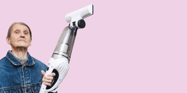 Proud elderly woman poses with a new handheld vacuum cleaner, the photo is isolated on a pink pastel background. - Foto, Bild
