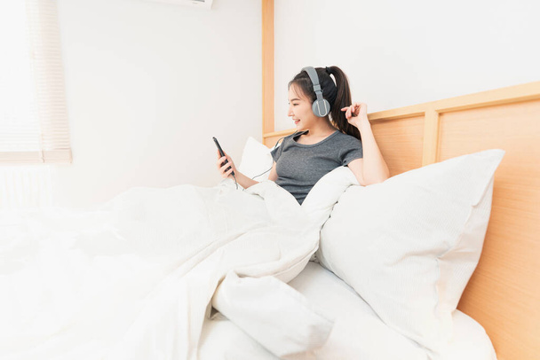 Υπνοδωμάτιο έννοια στο άνετο κρεβάτι ένα κορίτσι ακούγοντας τα αγαπημένα της τραγούδια φορώντας ένα ακουστικό το πρωί. - Φωτογραφία, εικόνα