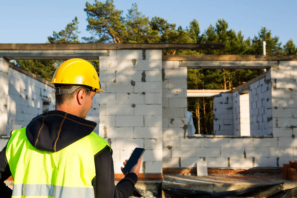 Construction worker talks on a smartphone in a yellow hardhat and reflective vest against the background of the construction of house-the walls and window openings are made of porous concrete blocks. - Photo, image