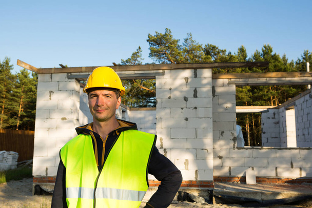 Project architect stands near construction site of a house with the walls are made of a porous concrete block and a brick. Construction worker in a protective yellow hardhat and a reflective vest. - Valokuva, kuva