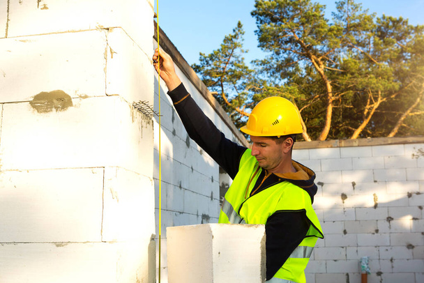 Construction worker at construction site measures the length of the window opening and the wall with tape measure. Cottage are made of porous concrete blocks, protective clothing - hardhat and a vest - Photo, image