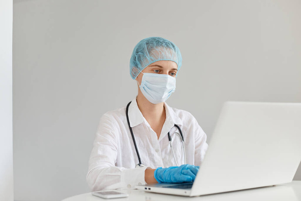 Indoor shot of concentrated woman doctor therapist wearing gown, surgical mask, medical cap and gloves, working in front of computer in clinic, having online colloquium. - Foto, Bild