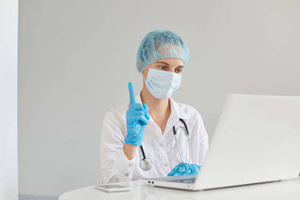Woman doctor therapist wearing gown, surgical mask, medical cap and gloves, working in front of computer in clinic, having idea for writing scientific medical article, pointing finger up. - Φωτογραφία, εικόνα