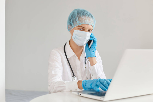 Portrait of serious woman doctor wearing gown, surgical mask, medical cap and gloves, posing in front of notebook, talking phone with college, working in hospital. - Foto, Bild