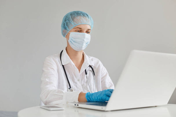 Caucasian woman doctor wearing gown, surgical mask, medical cap and gloves, posing in front of notebook, working working on a new scientific article. - Foto, afbeelding