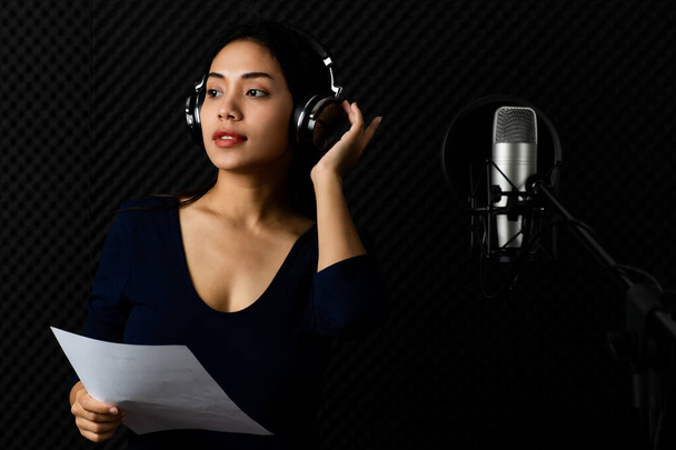 Szczęśliwa Asain kobieta jako piękna profesjonalna piosenkarka chwytająca słuchawki, uśmiech, cieszyć się ćwicząc atrakcyjny głos i śpiew występ do mikrofonu w ciemnym pokoju nagraniowym studio muzycznego - Zdjęcie, obraz