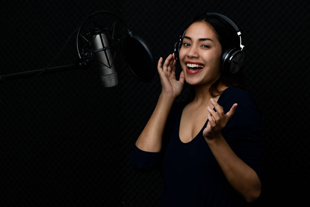 Mooie Aziatische vrouw als elegante professionele zangeres dragen van een koptelefoon en genieten van het uitvoeren van mooie lied zingen door het verhogen van zoete stem met vrolijke en emotionele opwinding in donkere muziek studio. - Foto, afbeelding