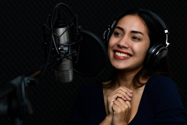 Csinos ázsiai nő, mint elegáns profi énekes visel fejhallgatót, és élvezze a gyönyörű dal éneklés emelésével édes hang boldog és érzelmi izgalom a sötét zenei stúdió. - Fotó, kép