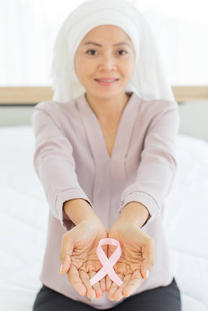 Cinta rosa mostró en la mano de la mujer asiática feliz, sentarse en la cama, sobrevivir del cáncer de mama, como símbolo que representa la conciencia internacional, el apoyo moral, dispuesto a luchar contra la enfermedad tumoral en el pecho - Foto, imagen