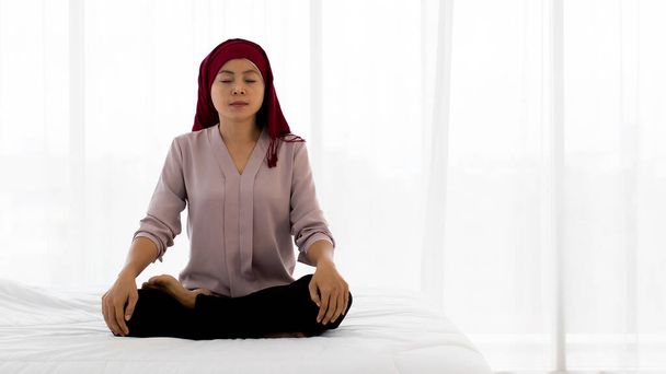乳がん患者としてのアジアの女性,赤いスカーフで頭をカバー,目を閉じて、足はリラックスして体を癒すために白いベッドの上に座っています,落ち着いて呼吸,そして静かな薬に簡単に瞑想 - 写真・画像