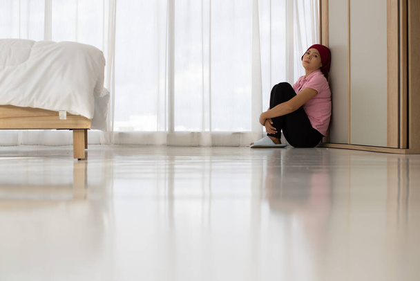 Lástima mujer asiática paciente de cáncer de mama tristemente sentarse solo en el suelo, apoyarse contra la pared, cerrar los ojos, y abrazar las rodillas en la esquina del dormitorio cerca de la cortina como desesperanzado y desalentado de la medicación fallida - Foto, imagen