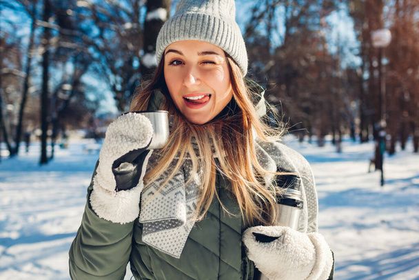 Happy woman having hot tea out of thermos in winter park. Drinks to warm up in snowy frosty weather outdoors during walk. Young woman showing tongue holding cup wearing warm clothes - Фото, изображение