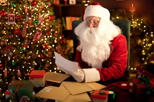 Santa Clause is prepares gifts for children for Xmas at his desk at home while reading wish lists - Fotoğraf, Görsel