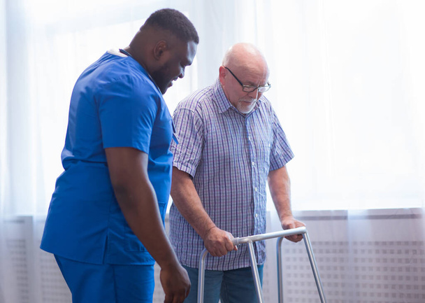 Caregiver is teaching old man to walk with walker. Professional nurse and patient in a nursing home. Assistance, rehabilitation and health. - Foto, immagini