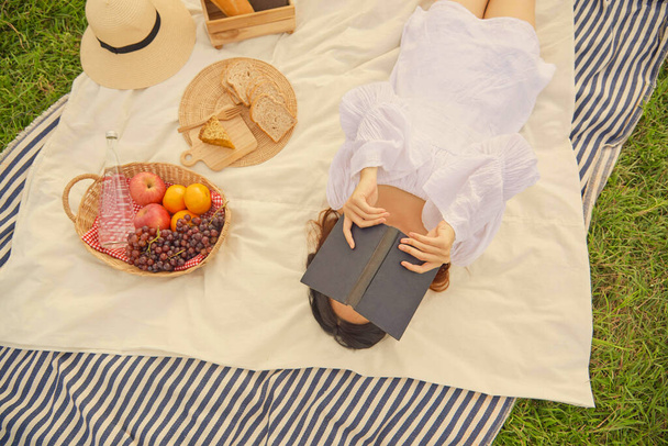 Концепция пикника Женщина в великолепном белом платье спит на белой и синей полосатой ткани с черной книгой покрывая лицо, находясь на хороший пикник. - Фото, изображение