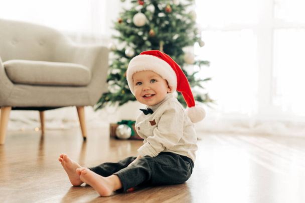 A laughing boy in a Santa Claus hat sits on the floor against the background of a Christmas tree - Фото, изображение