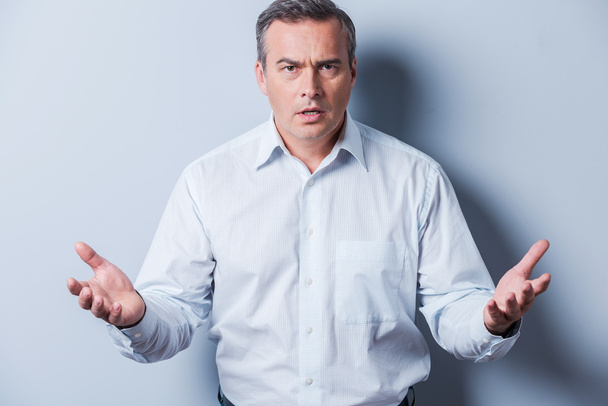 Can you believe it? Frustrated mature man in shirt looking at camera and gesturing while standing against grey background - Fotó, kép