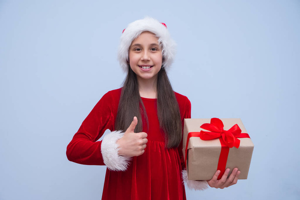 Smiling teenage brunette girl with long hair in red Santa outfit in her hands holds box with gift for Christmas holidays and New Year looks at the camera shows the class cool. Place for advertising - Photo, Image