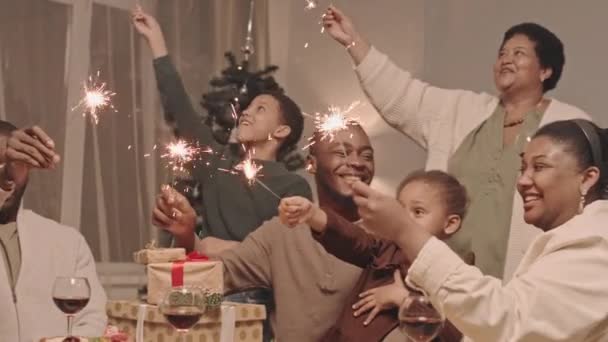 Medio tiempo de alegres miembros de la familia afroamericana de diferentes generaciones sentados a la mesa, sosteniendo luces encendidas de Bengala y divirtiéndose en Navidad - Imágenes, Vídeo