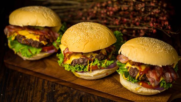 Three hamburgers with beef burger cutlet, fried onion, spinach, ketchup sauce and cheese, served on wood chopping board over dark wooden background - Photo, Image