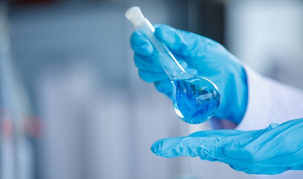 Zárja be a laboratóriumi főzőpoharat kék folyadékkal az elemző kezében, műanyag kesztyű elmosódott háttérrel. Koncepciókutatás - Fotó, kép
