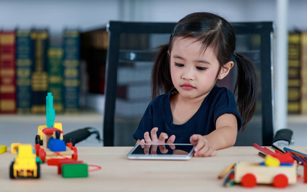 Small Asian pigtails hairstyle preschooler kindergarten girl sitting at table full of plastic wood truck toys smiling look at camera touching tablet computer in living room at home in front bookshelf. - Photo, Image