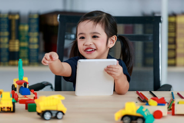 Małe azjatyckie warkocze fryzura przedszkolak dziewczyna siedzi przy stole pełnym plastikowych zabawek ciężarówka uśmiechnięty spojrzeć na aparat dotykając tabletu w salonie w domu w przedniej półce książki. - Zdjęcie, obraz