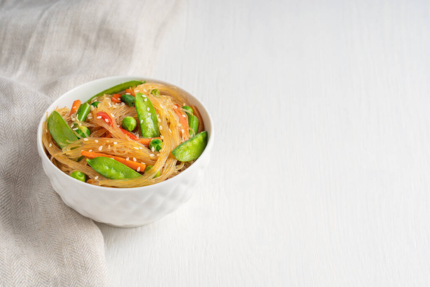 Japchae savory vegetarian korean popular dish of stir-fried glass noodles and vegetables such as green peas, bean and carrot served in bowl with textile on white wooden table. Image with copy space - Foto, Imagen
