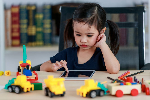 Little cute happy Asian pigtails hairstyle kindergarten girl sitting posing at table full of plastic wood truck toys in living room in front bookshelf. - Foto, Imagem