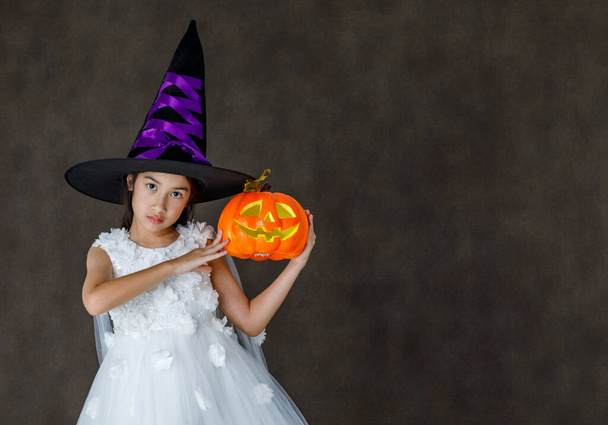 Portrait close up studio shot of Asian cute little girl in witch dress costume with tall hat stand smile look at camera hold orange pumpkin in hand ready to play trick or treat on Halloween festival. - Foto, Imagen