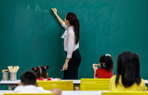Portrait shot of Asian young beautiful female English language professor teacher lecturer standing using chalk writing alphabet letters question on chalkboard for students in primary school classroom. - Photo, Image