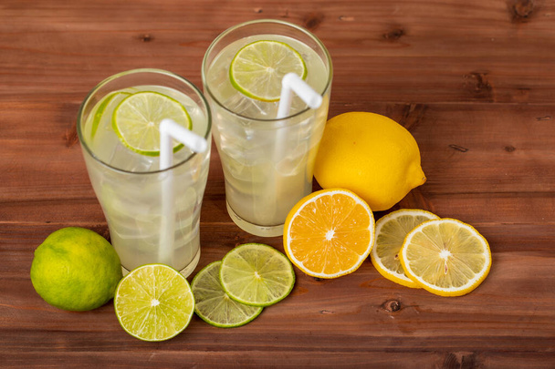 Dwa orzeźwiające kieliszki zimnego soku owocowego ze słomką, lodową limonką i sokiem z cytryny, ozdobione świeżą limonką i cytryną na drewnianym stole. Pojęcie odświeżania. - Zdjęcie, obraz