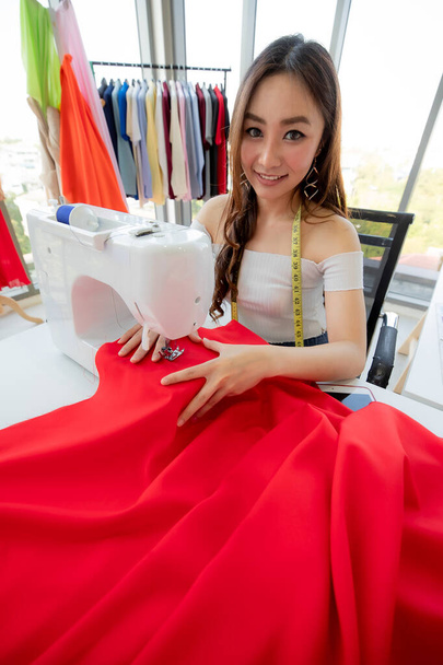 Šťastná asijská švadlena s měřicí páskou na krku práce v ateliéru a šití červené tkaniny se strojem - Fotografie, Obrázek