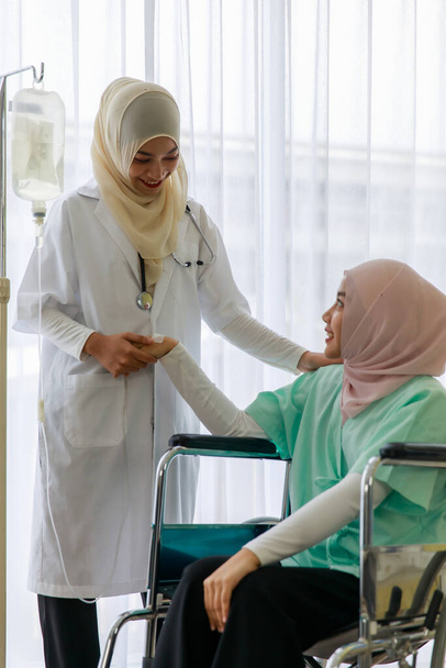 Giovane medico musulmano femminile parlare e rallegrare giovane pateint musulmano malato seduto sulla sedia a rotelle in ospedale. - Foto, immagini