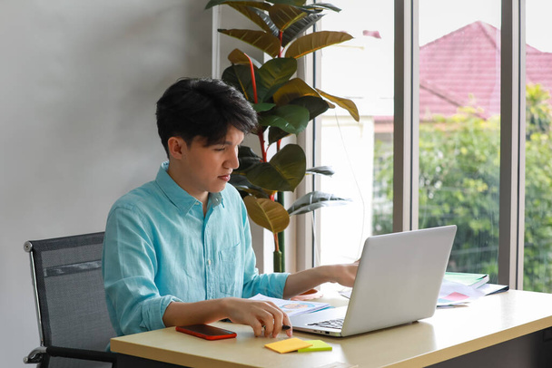 Young handsome friendly asian male employee sit on black chair work with silver laptop on desk full of documents mobile phone and post it note in glass room office with beautiful tree in background. - Photo, Image