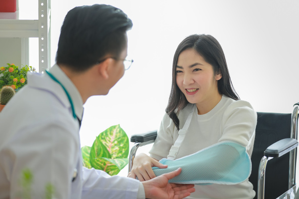 Crop médico étnico que explica joven mujer asiática cómo usar el cabestrillo del brazo después de un trauma de trabajo en el hospital - Foto, imagen