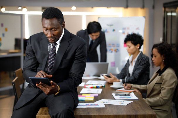 Africké černošky výkonný úspěšný muž majitel podnikání sedí za stolem v kryté kanceláři s pozadím zaměstnance nebo kolegy, pomocí tablet seriózní analýzy a plánování práce s koncentrací - Fotografie, Obrázek