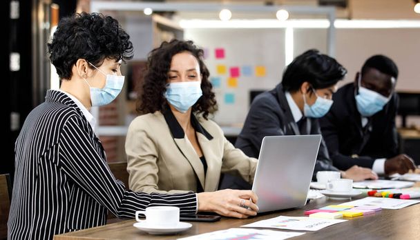 Wielorasowe inteligentne formalne biznesmeni siedzą twarzą w twarz ze spotkaniem i prowadzą dyskusję grupową w biurze, mając maski na twarzy jako nową normę w celu ochrony lub zapobiegania wirusowi w czasie pandemii - Zdjęcie, obraz
