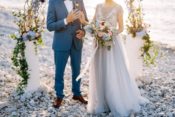 Kotor, Černá Hora - 08.06.17: Nevěsta v krajkových šatech dává zásnubní prsten na prst ženicha v modrém obleku. Novomanželé stojí pod svatebním obloukem na pláži. Zavřít - Fotografie, Obrázek