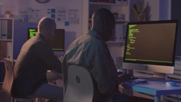 コンピュータのモニターの前に机に座っている2人の男性多民族のプログラマーの後方ビューの遅いショット夜に暗いオフィスに滞在する新しいソフトウェアを開発するプログラムコードを書く - 映像、動画
