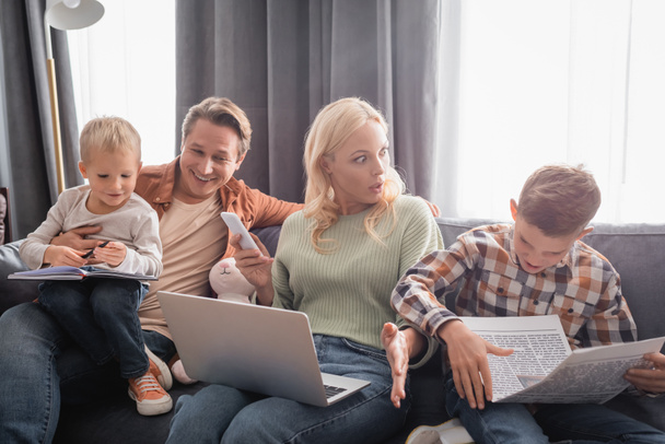 shocked and busy woman with laptop and smartphone working near cheerful family having fun on couch - Photo, Image