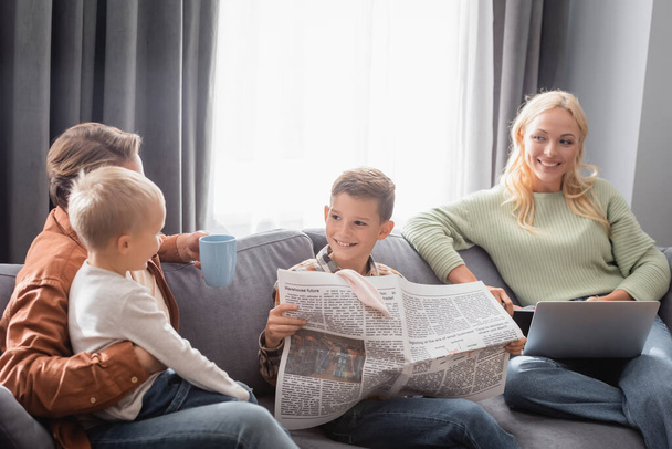 cheerful boy holding newspaper near family on couch in living room - Фото, изображение
