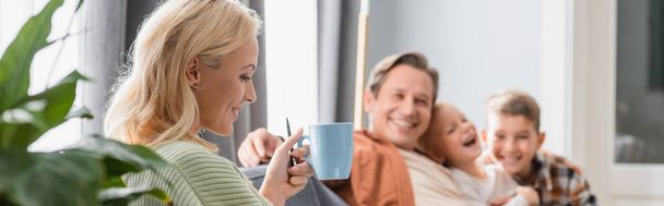 happy woman drinking tea near blurred family in living room, banner - Photo, Image
