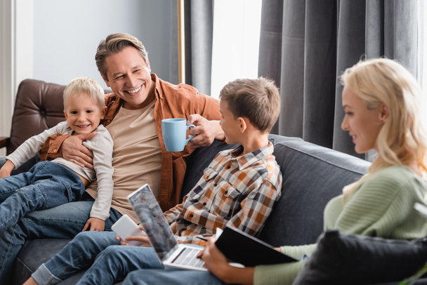 happy man with cup of tea sitting on sofa with sons while blurred wife working on laptop - Photo, Image