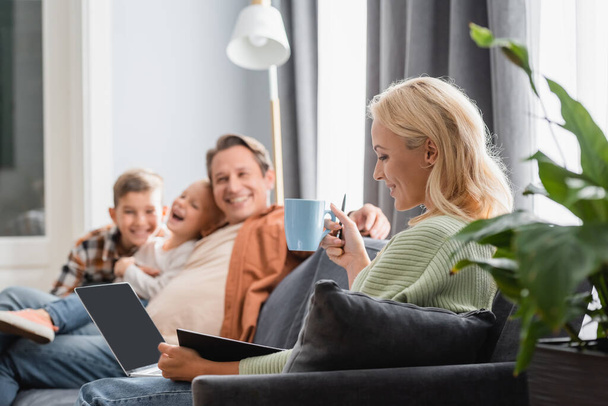 smiling woman with cup of tea working with laptop and notebook near blurred family on sofa - Photo, Image