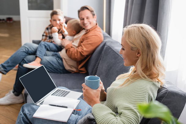 woman with notebook, laptop and cup of tea sitting on couch near blurred cheerful family - Fotoğraf, Görsel