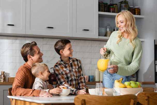 веселая женщина наливает апельсиновый сок во время завтрака с мужем и детьми - Фото, изображение