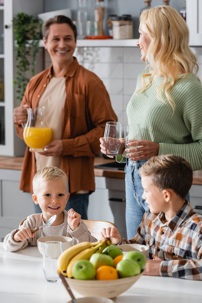 smiling parents looking at each other while holding glasses and orange juice near sons having breakfast - Fotoğraf, Görsel