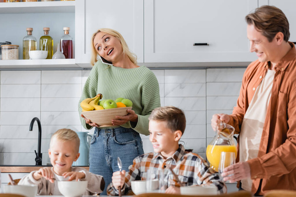 woman with bowl of fresh fruits talking on smartphone near man with orange juice ands boys having breakfast - Foto, afbeelding